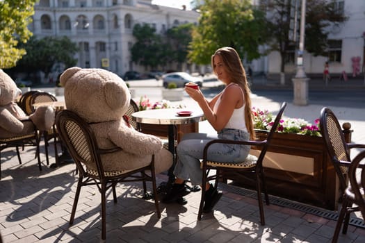 A woman sits cafe with a teddy bear next to her. The scene is set in a city with several chairs and tables around her. The woman is enjoying her time at the outdoor cafe