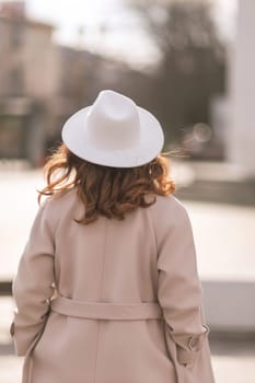 Back view of happy woman wearing hat and coat walking down street on sunny spring day. People, lifestyle, travel and vacations concept.