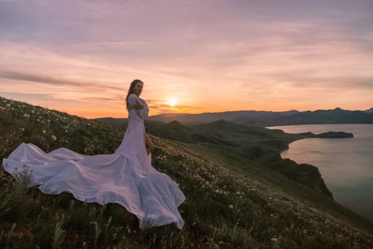 A woman in a white dress stands on a hill overlooking a body of water. The sky is a mix of orange and pink hues, creating a serene and romantic atmosphere. The woman's dress billows in the wind