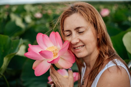A woman is holding a pink lotus flower and smiling. The flower is surrounded by green leaves, and the woman is standing in a field of flowers