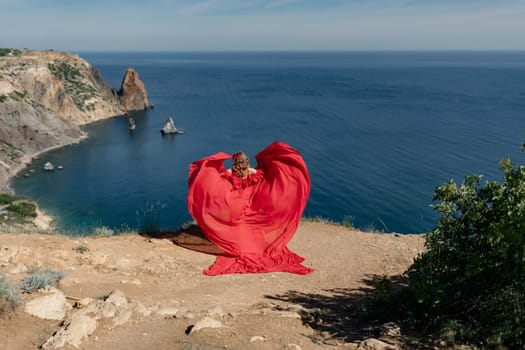 A woman in a red dress is standing on a rocky beach, with the ocean in the background. She is wearing a red dress that is flowing out behind her, creating a heart shape. The scene is serene