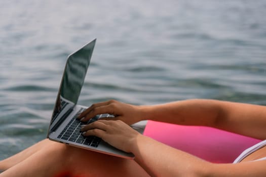 A woman is typing on a laptop while laying on a pink inflatable raft in the water. Concept of relaxation and leisure, as the woman enjoys her time by the water while working on her laptop