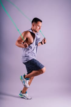 A man with a grey tank top is holding a green jump rope with his elbow bent and waist twisted, showing off his toned chest and wrist strength