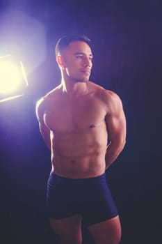 A man with a bare chest is posing against a dark backdrop, showing off his muscular physique. His head, hands, stomach, arms, shoulders, legs, and neck are visible in the flash photography