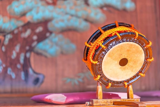 Japanese shime-daiko drum whose edges are laced with thick orange rope placed on a wooden terendai support with bachi sticks next to a zabuton cushion in front of a giant mural depicting a pine tree.