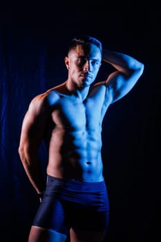 A man with a bare chest is posing against a dark backdrop, showing off his muscular physique. His head, hands, stomach, arms, shoulders, legs, and neck are visible in the flash photography