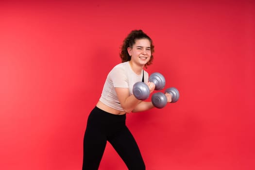 Teenage sportive girl exercises with dumbbells to develop muscles isolated on a red background.