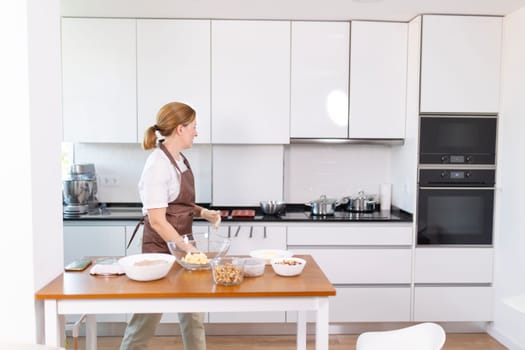 A woman is cooking in a kitchen with a white countertop and white cabinets. She is wearing an apron and has a smile on her face