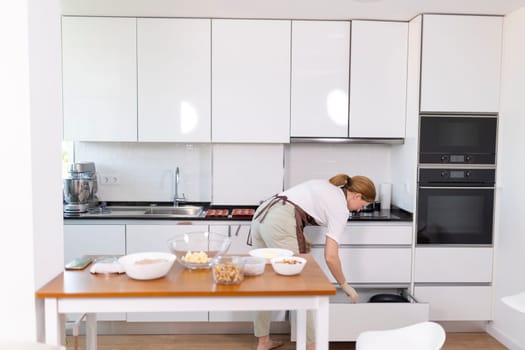 A woman is in a kitchen, preparing food. The kitchen is clean and well-organized, with a wooden table and chairs. The woman is wearing an apron and she is focused on her task