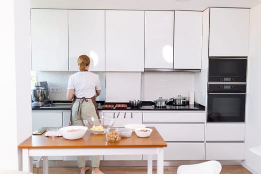 A woman is cooking in a kitchen with white cabinets and a wooden table. The kitchen is well-equipped with a microwave, oven, and toaster. The woman is wearing an apron and she is preparing a meal