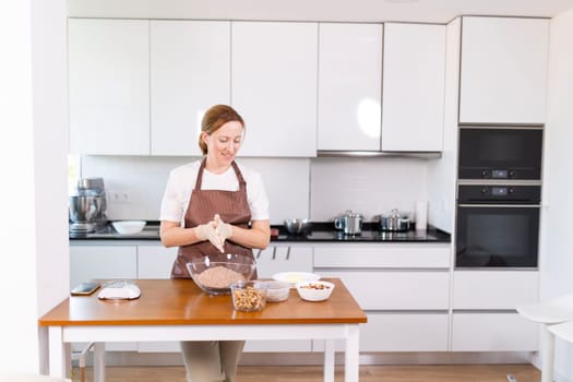 A woman is standing in a kitchen, preparing food. She is wearing an apron and gloves, and there are several bowls and cups on the table. The kitchen is well-equipped with an oven, microwave