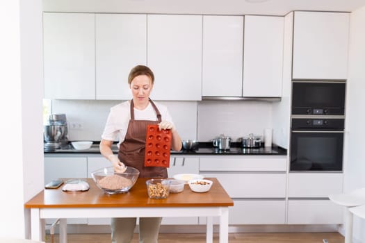 A woman is in a kitchen preparing food. She is wearing an apron and standing at a wooden table. The kitchen is well-equipped with appliances such as an oven, microwave, and toaster