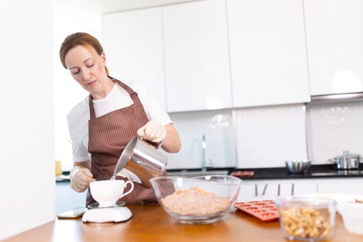 A woman is pouring a liquid into a cup while wearing an apron. The kitchen is clean and well-organized, with various items such as bowls, cups, and a scale. The woman is focused on her task