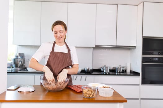 A woman is making a dessert in a kitchen. She is wearing an apron and gloves. The kitchen is well-equipped with appliances such as an oven, microwave, and toaster