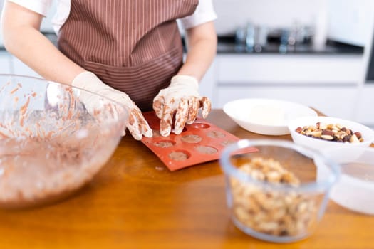 A woman is making cookies on a wooden table. She is wearing an apron and gloves. There are several bowls and cups on the table, and a few spoons. The woman is using a cookie cutter to make cookies