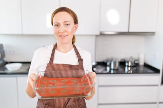 A woman is holding a glass baking dish with cookies in it. She is wearing an apron and smiling