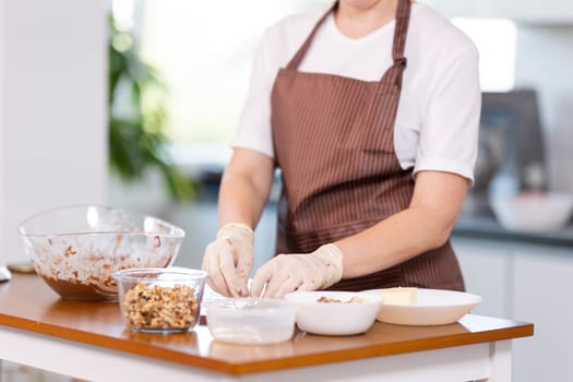 A woman is preparing food on a wooden table. She is wearing an apron and gloves. There are several bowls and spoons on the table