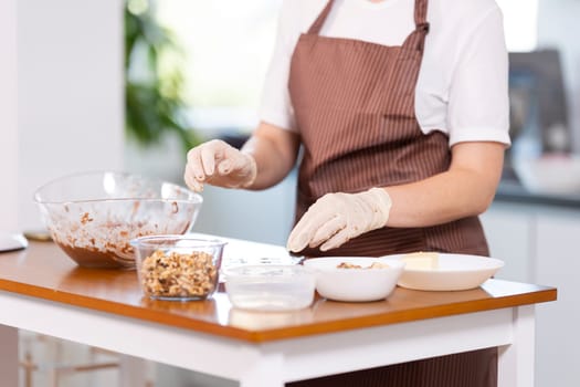 A woman is preparing food on a table with a bowl of nuts and a bowl of chocolate. She is wearing gloves and an apron