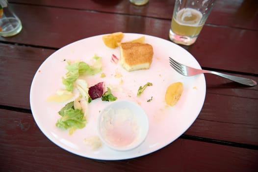 Leftover food on a white plate on wooden table, close-up. Food and drink consumerism. Top view