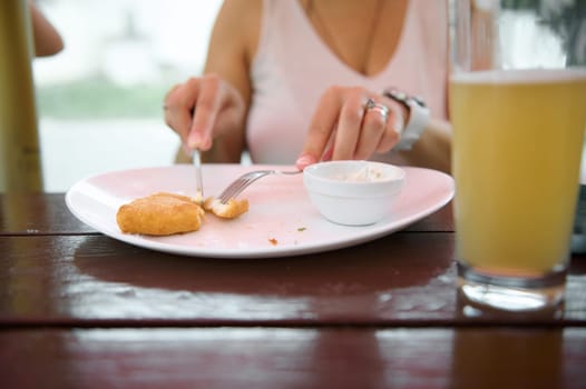 Close-up woman at restaurant, having lunch. Female hands using fork and kitchen knife, cut fried cheese. A cup of beer on table on foreground. Woman eating in cafe outdoors. Food and drink consumerism