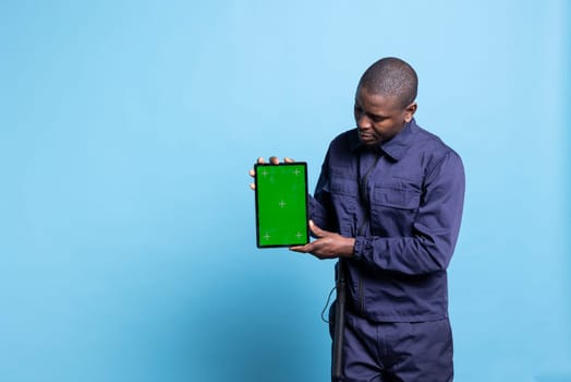 Trained armed bodyguard holds a tablet with greenscreen layout on camera, presenting isolated copyspace against blue background. Security officer posing in uniform with device in studio.