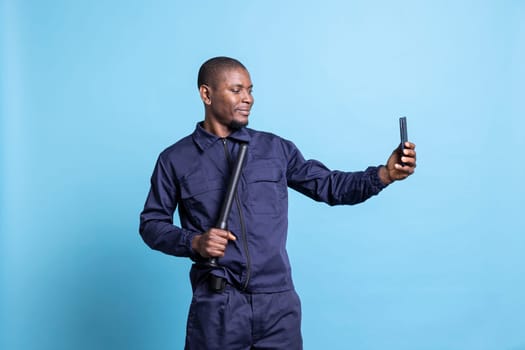 Smiling security agent taking photos with his baton on camera, using a smartphone to take pictures against blue background. Young guard makes memories in his patrol uniform in studio.
