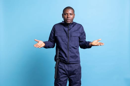 Clueless unsure security officer shrugging against blue background, raising his shoulders and showing i dont know sign. Bodyguard acting uncertain and doubtful, confused personnel.