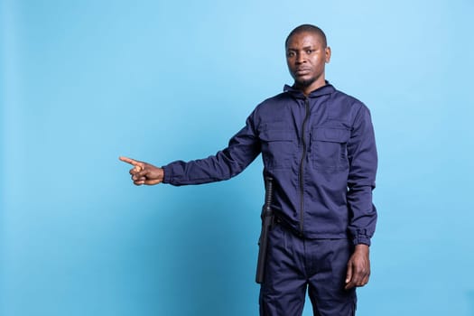 African american bodyguard pointing to something aside in studio, working on an advertisement against blue background. Male security officer in uniform indicates a left or right direction.