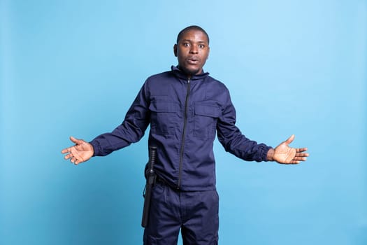 Security guard in uniform is acting surprised in front of the camera, being in shock after finding out something. African american protection agent with baton feels amazed against blue background.