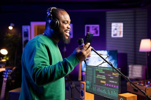 Pleased artist does a recording session in his home studio with professional gear, performing a new song on microphone. Smiling composer reading the lyrics and notes on smartphone.