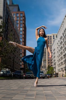 Beautiful Asian ballerina in blue dress posing in splits outdoors. Urban landscape. Vertical photo