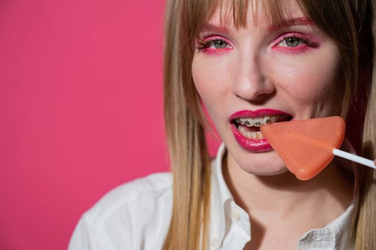 Portrait of a young woman with braces and bright makeup eating a lollipop on a pink background