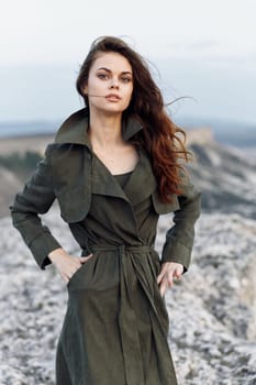 Confident woman in trench coat standing on rugged hillside with hands on hips