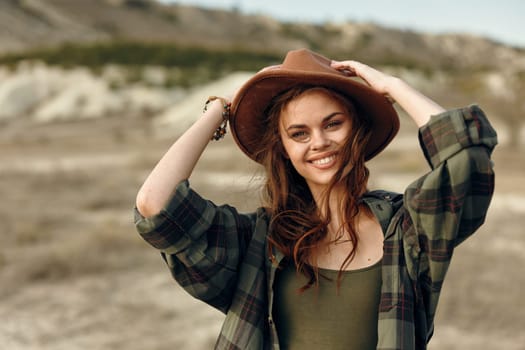 Stylish woman in plaid shirt and hat poses with hands on head in vast desert landscape