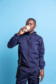 African american officer drinking a cup of coffee in studio, serving caffeine beverage to get more energy after a sleepless night shift. Patrolling employee agent poses in front of camera.