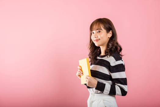 Portrait of beautiful Asian young woman teenage smiling holding a yellow book and reading, studio shot isolated on pink background with copy space