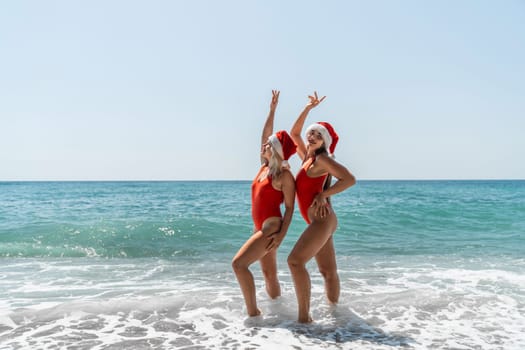 Women Santa hats ocean play. Seaside, beach daytime, enjoying beach fun. Two women in red swimsuits and Santa hats are enjoying themselves in the ocean waves and raising their hands up