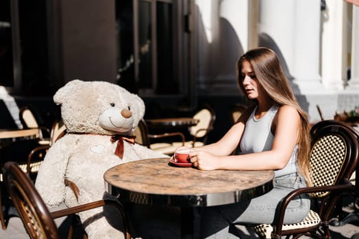 A woman sits cafe with a teddy bear next to her. The scene is set in a city with several chairs and tables around her. The woman is enjoying her time at the outdoor cafe