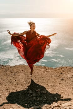 Woman red dress sea. Female dancer in a long red dress posing on a beach with rocks on sunny day. Girl on the nature on blue sky background