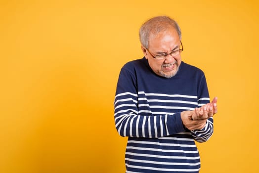 Portrait Asian old man with glasses holding his painful hand studio shot isolated yellow background, senior man pensioner feeling pain hand pain wrist bone joint problem