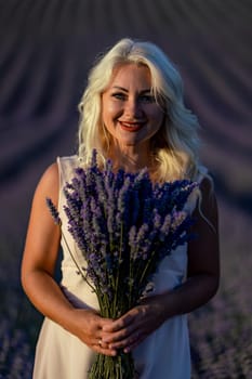 Blonde woman poses in lavender field at sunset. Happy woman in white dress holds lavender bouquet. Aromatherapy concept, lavender oil, photo session in lavender.