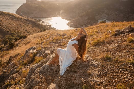 A woman in a white dress is sitting on a rock overlooking a body of water. She is enjoying the view and taking in the scenery