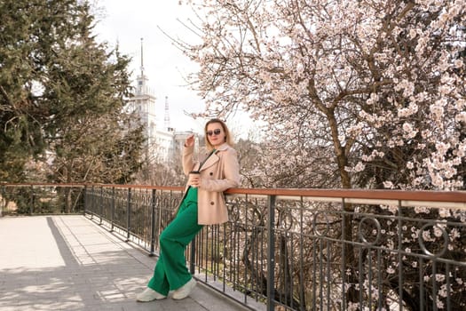 A woman is standing on a ledge with a cup of coffee in her hand. She is wearing sunglasses and a tan coat. The scene is set in a park with cherry blossoms in bloom. The woman is enjoying the view