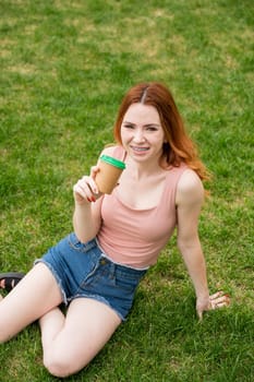 A beautiful young woman with braces on her teeth drinks from a cardboard cup while sitting on the grass. Vertical photo