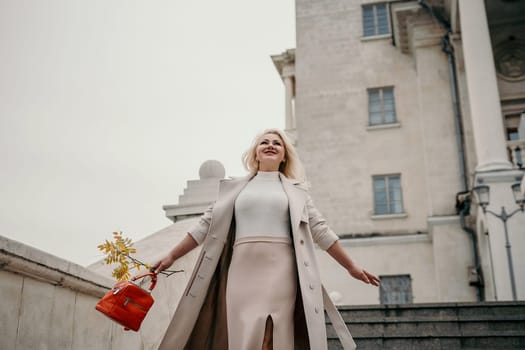 A woman wearing a white coat and holding a red purse is standing on a staircase. She is smiling and she is happy