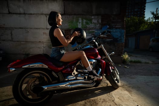 Brunette woman in denim shorts puts on a helmet while sitting on a red motorcycle