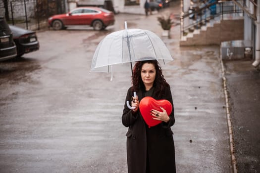 A woman holding a red heart under a clear umbrella. The umbrella is open and the woman is standing in the rain