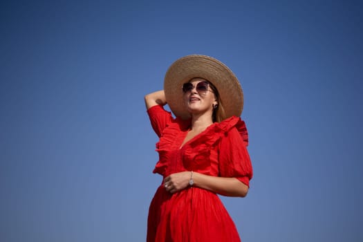 A woman in a red dress and straw hat is smiling and looking up at the sky. Concept of happiness and relaxation, as the woman is enjoying her time outdoors