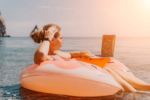 Woman freelancer works on laptop swimming in sea on pink inflatable ring. Pretty lady typing on computer while floating in the sea on inflatable donut at sunset. Freelance, remote work on vacation