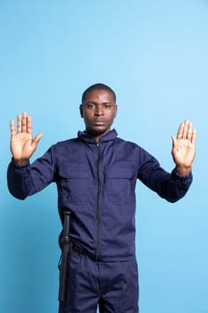 Male security officer putting his hands up as a stop sign on camera, showing a limitation symbol against blue background. Serious patrol agent restricting the access, does a rejection gesture.